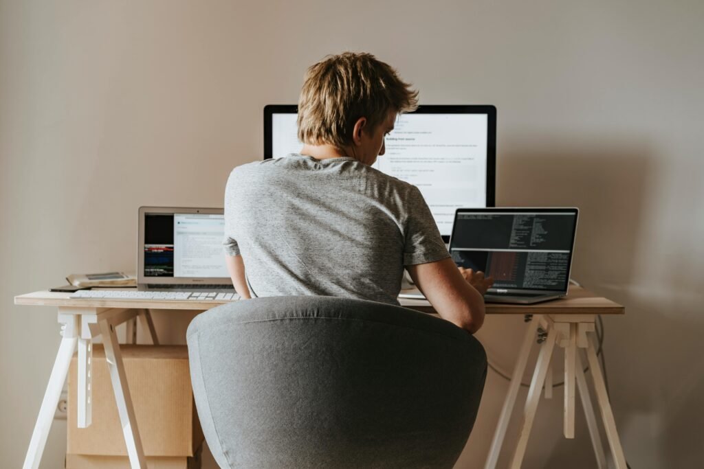 pexels-photo-4974907-4974907 Young man coding at home office with multiple laptops, representing remote work and technology.